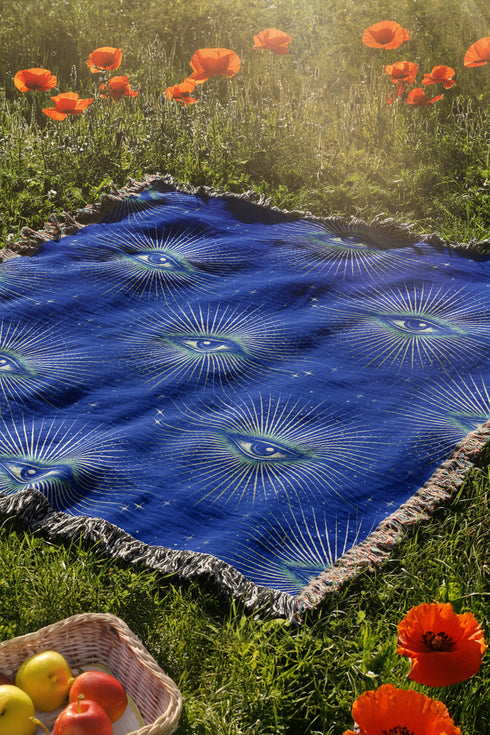 Woven cotton throw blanket with a deep blue background featuring a repeated pattern of all-seeing eyes in gold and seafoam green. The blanket has a colorful fringed edge.